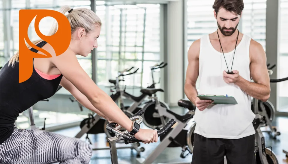 Athletin auf dem Velo-Ergometer, ein Trainer notiert die Werte auf dem Tablet